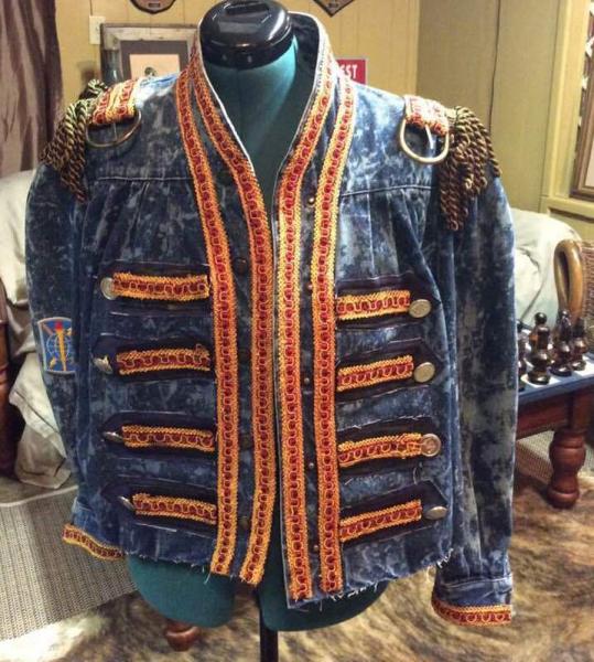 20203 Michael J Bleached Denim Jacket has added leather, buttons, and cording. Epaulets of fringe with belt buckles are added. Sizes range from small to large.
